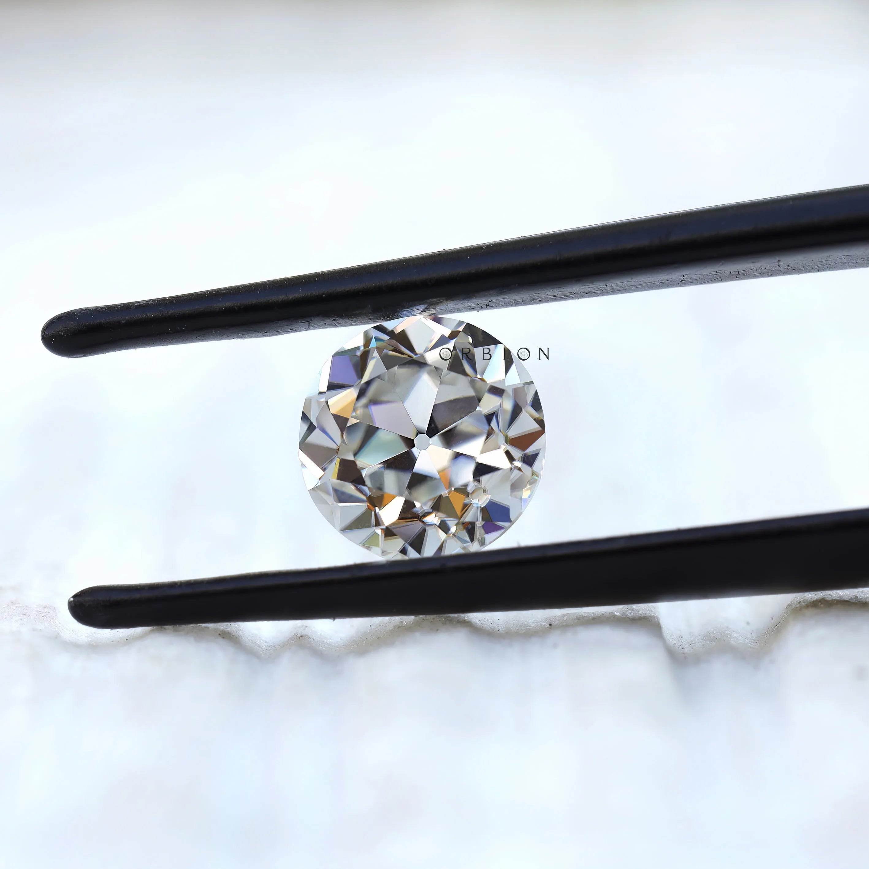 Round old European cut moissanite held in black tweezers