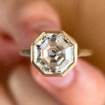 Moissanite octagon step cut bezel set cathedral style solitaire ring in 10K yellow gold.