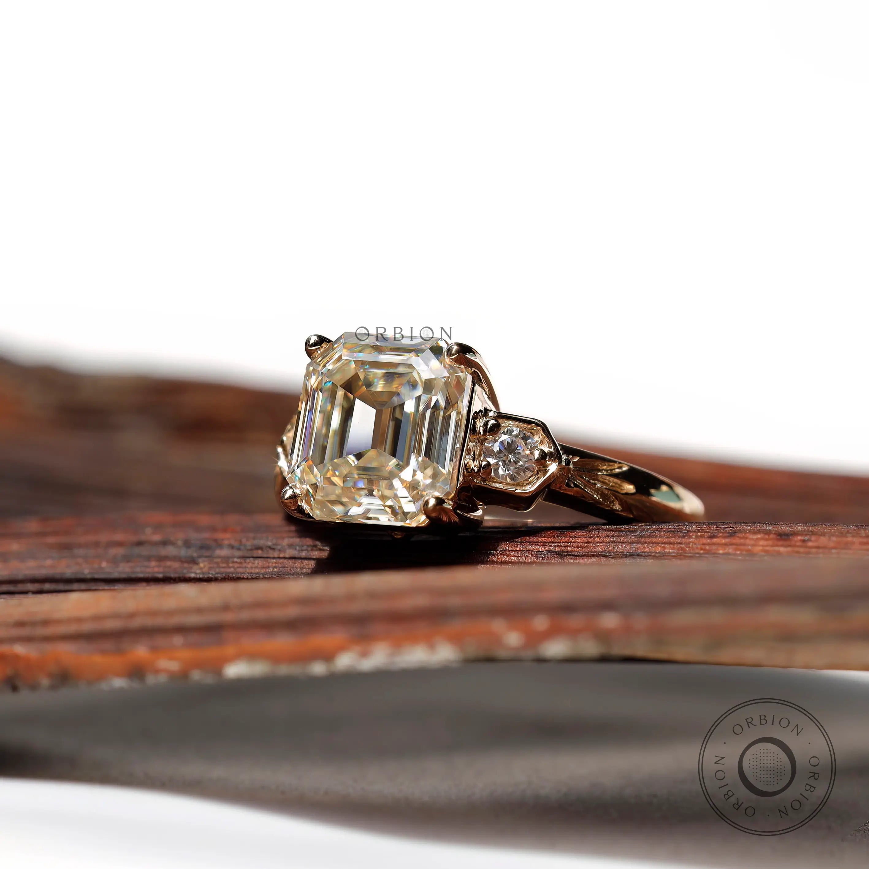 Profile shot of the three-stone engagement ring, highlighting the Asscher cut Moissanite and two round side stones in 10K yellow gold.