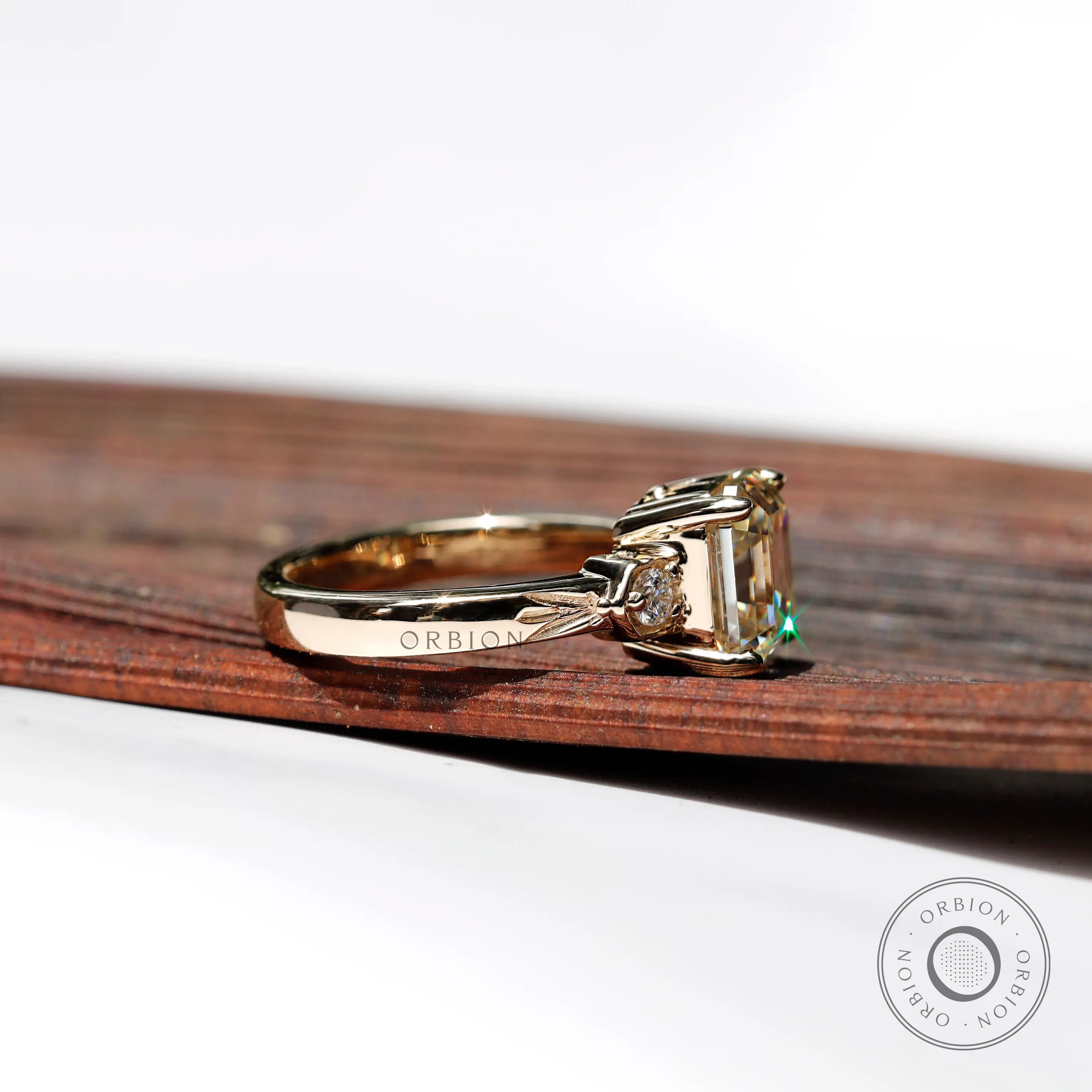 Side view of a three-stone engagement ring with a step-cut Asscher Moissanite center and two round Moissanite side stones, set in 10K yellow gold.