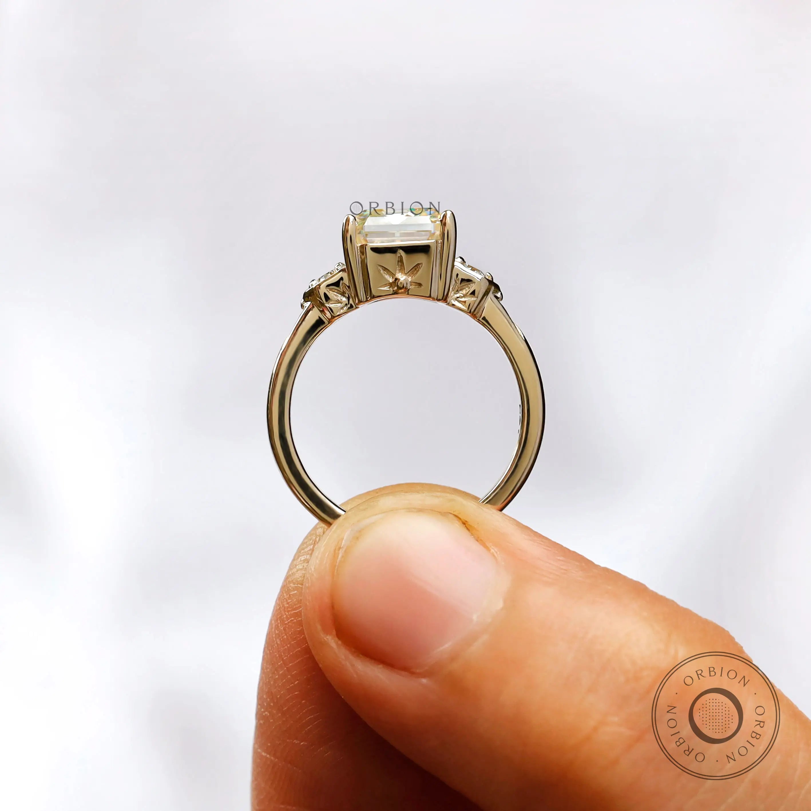 Hand-worn three-stone engagement ring with an Asscher cut Moissanite center stone and round Moissanite side stones in 10K yellow gold.