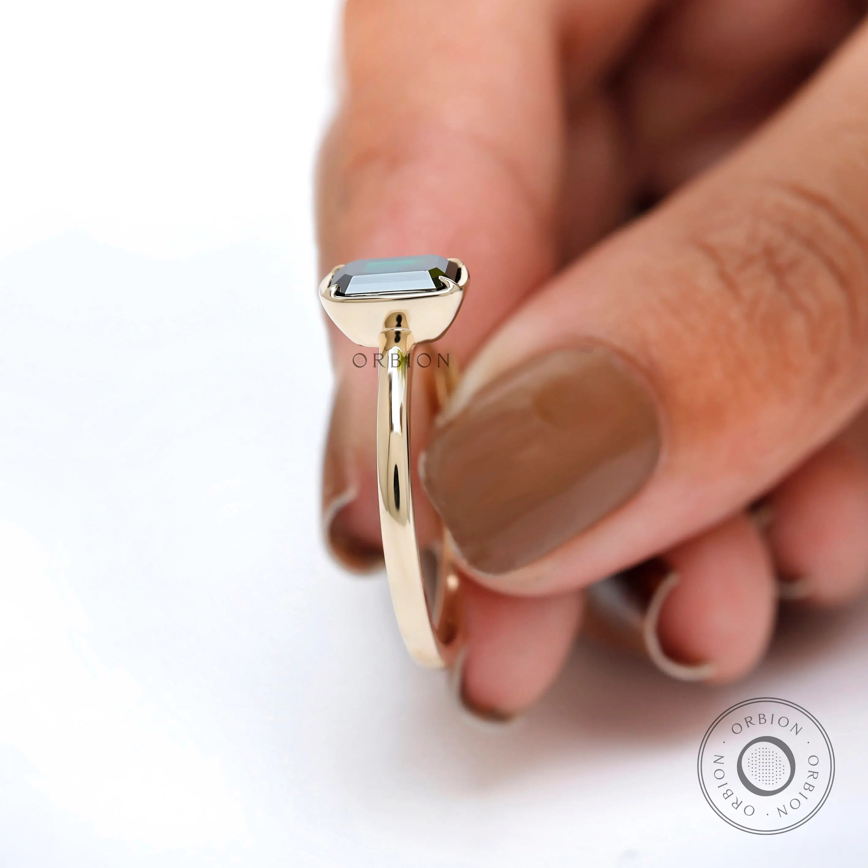 Bezel set emerald cut moissanite ring held between fingers.