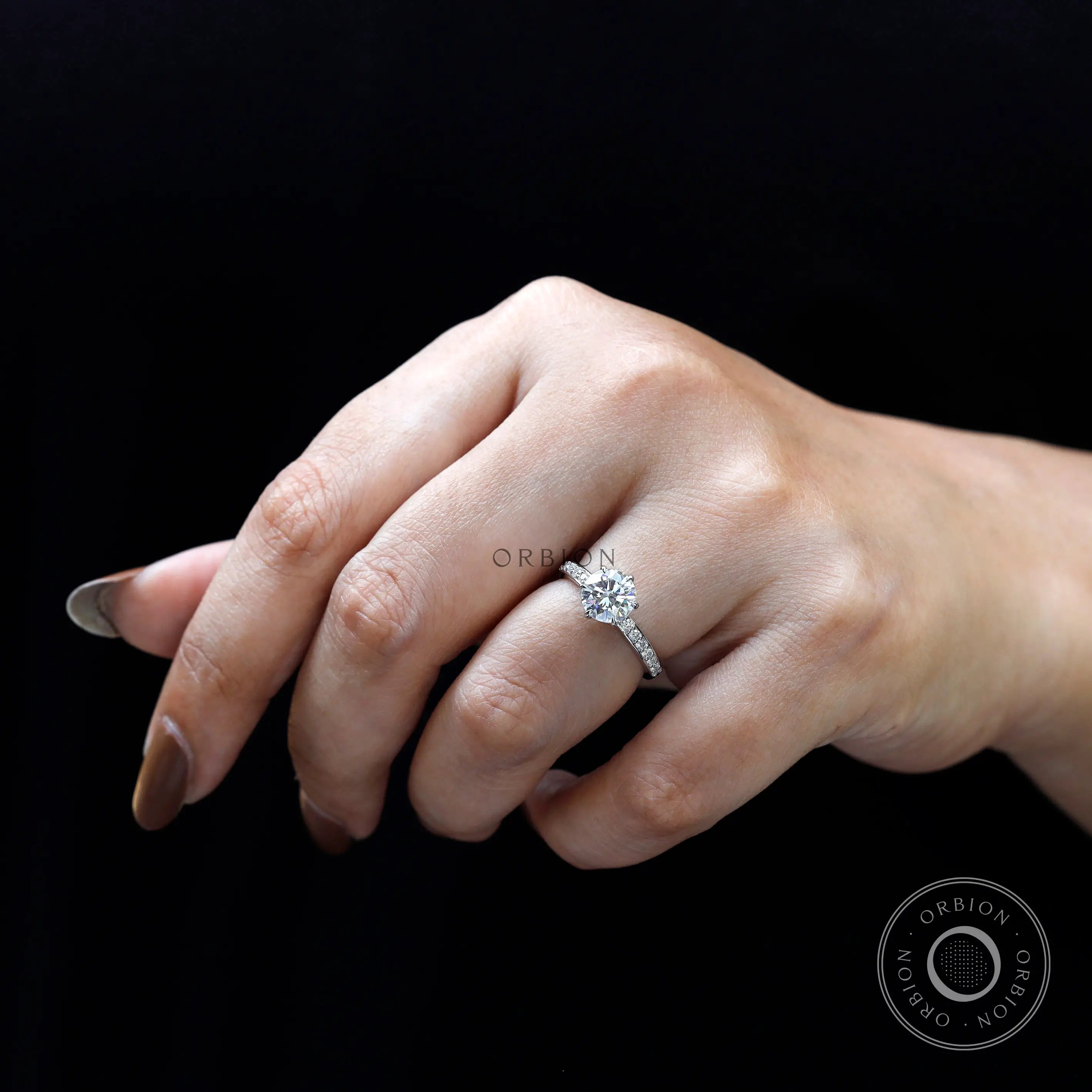Round moissanite solitaire engagement ring worn on hand against black background.