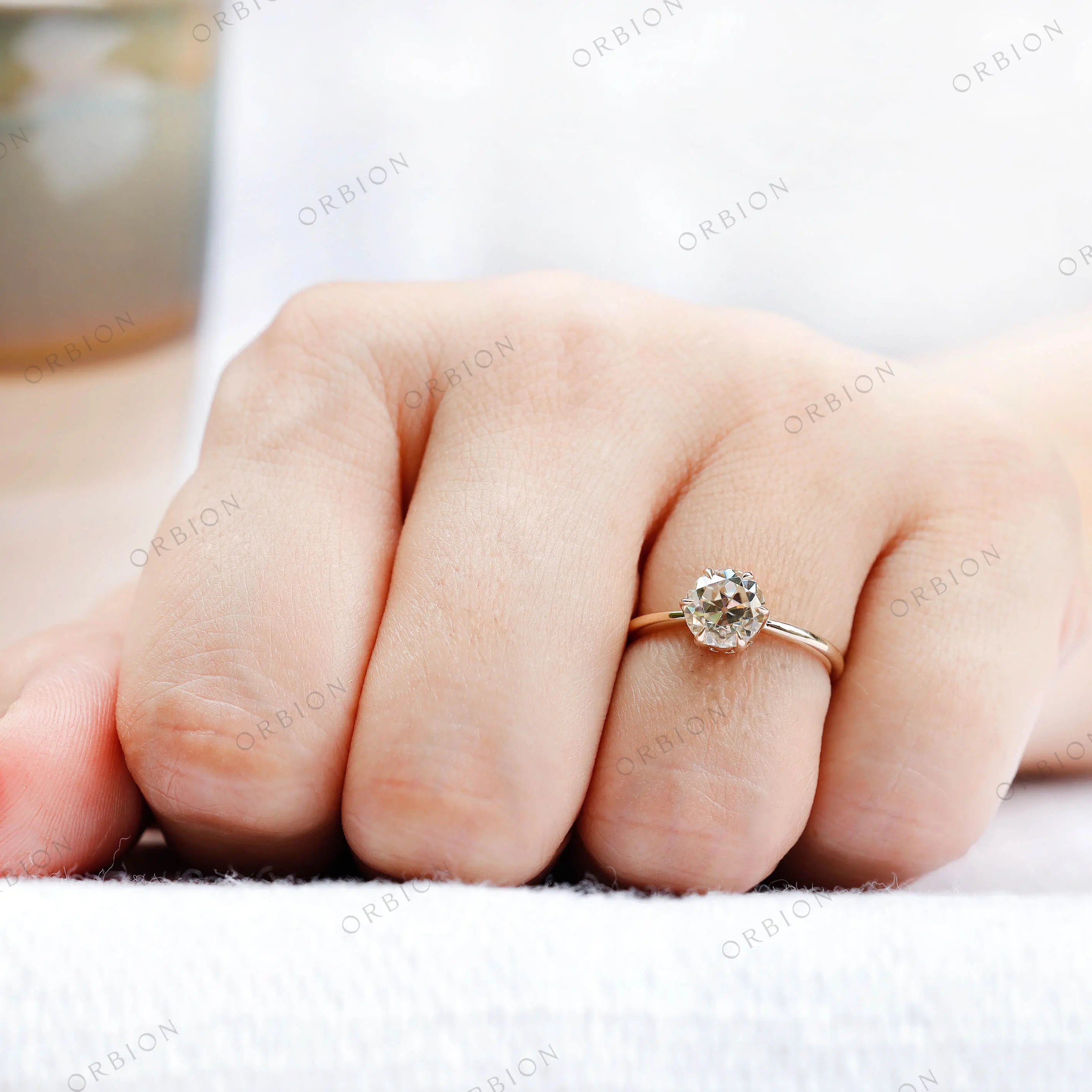 Close-up of the solitaire engagement ring being held, highlighting the round moissanite center stone in 10K yellow gold.